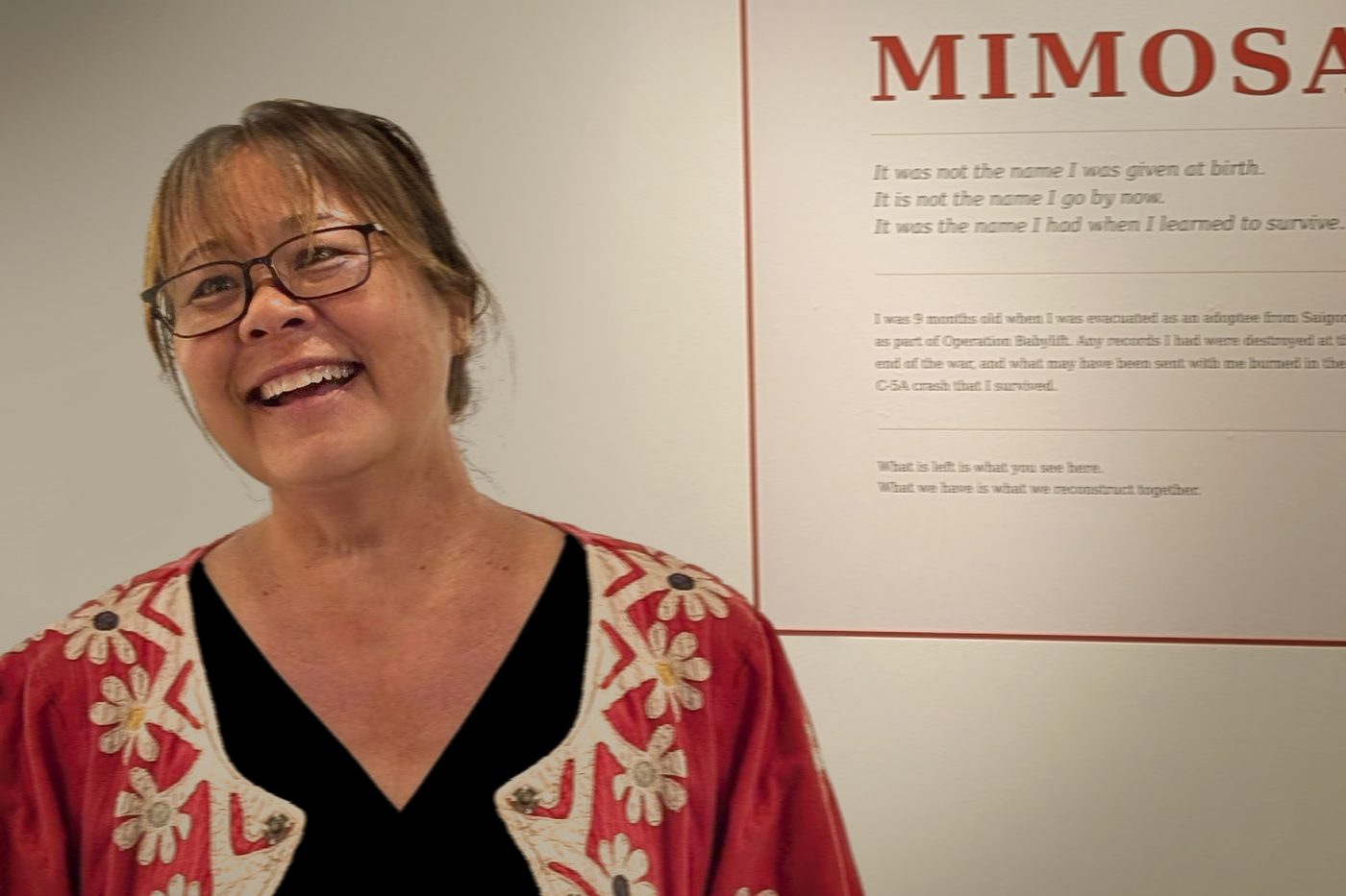 Devaki smiling beside a gallery wall panel titled MIMOSA at the exhibition opening